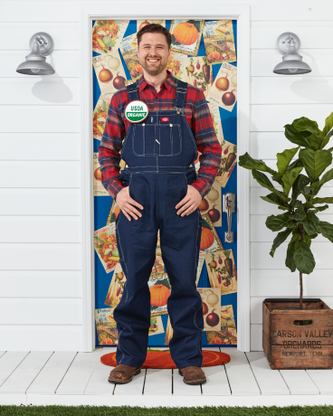 muž v kostýmu farmáře s kombinézou, kostkovanou košilí, organickým knoflíkem usda stojící přede dveřmi zdobenými na halloween