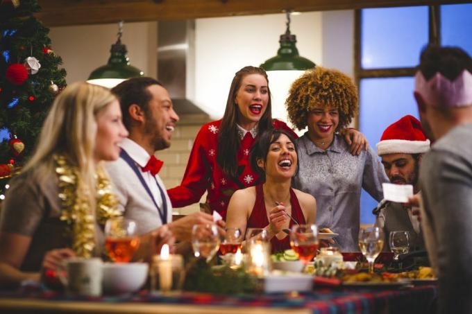 skupina přátel, kteří se smějí, když sedí kolem stolu, jedí a popíjejí o Vánocích jeden muž čte vtip z vánočního sušenky, nosí se klobouky a vánoční svetry