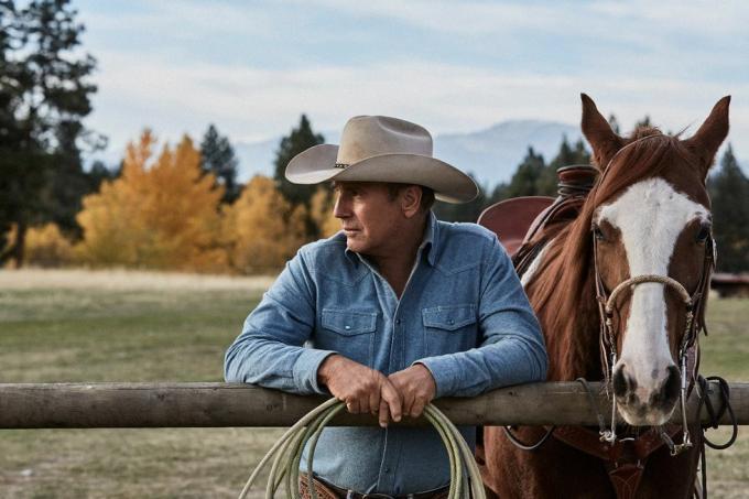 Kevin costner v Yellowstone vedle koně opírajícího se o plot s provazem v rukou na sobě vybledlou modrou džínovou košili a béžový kovbojský klobouk