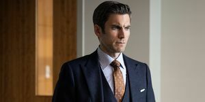 wes bentley v tmavě šedém třídílném obleku s hnědou potištěnou kravatou a bílou a modrou pruhovanou košilí stojící v kanceláři s rukama v kapsách