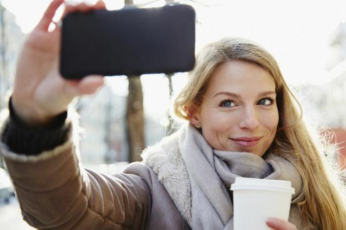 pohledná žena, která v zimě chodí po ulicích města, dělá si selfie a drží kávu, aby šla, podsvícená