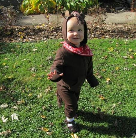 dítě oblečené jako kůň v hnědé mikině s kapucí a kalhotách s červeným šátkem kolem krku má kapuci hřívu z příze a hnědé plstěné uši