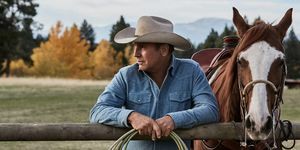 Kevin costner v Yellowstone vedle koně opírajícího se o plot s provazem v rukou na sobě vybledlou modrou džínovou košili a béžový kovbojský klobouk