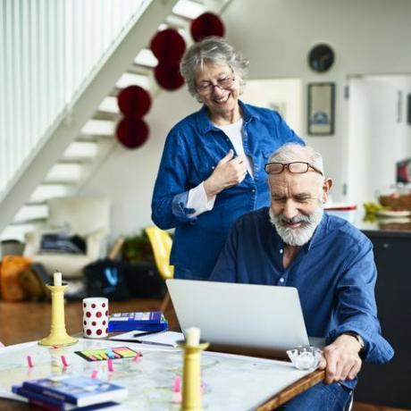 starší žena s úsměvem a dívá se přes rameno manžela, když zkoumá a čte online recenze o místech k pobytu a místech k návštěvě, poznámkách a průvodcích na stole, rezervaci dovolené online