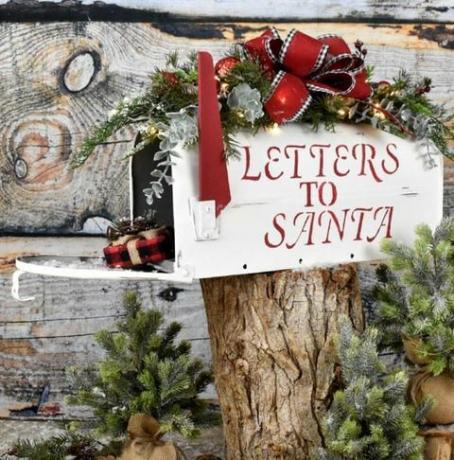 bílá poštovní schránka s červenými písmeny "dopisy Ježíškovi", sedící na kládě, dveře je otevřený a vylévá se z něj dárek, schránka je zakončena vánoční zelení a červenou luk